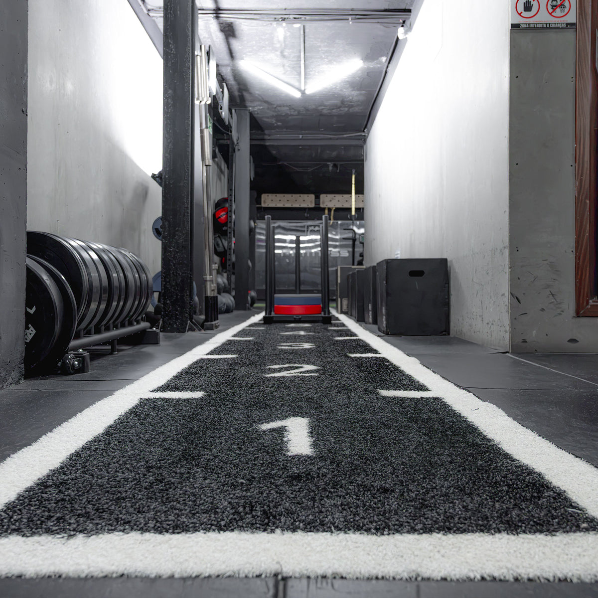 Pista de relva preta numerada A038-BL instalada num ginásio, com trenó de treino ao fundo